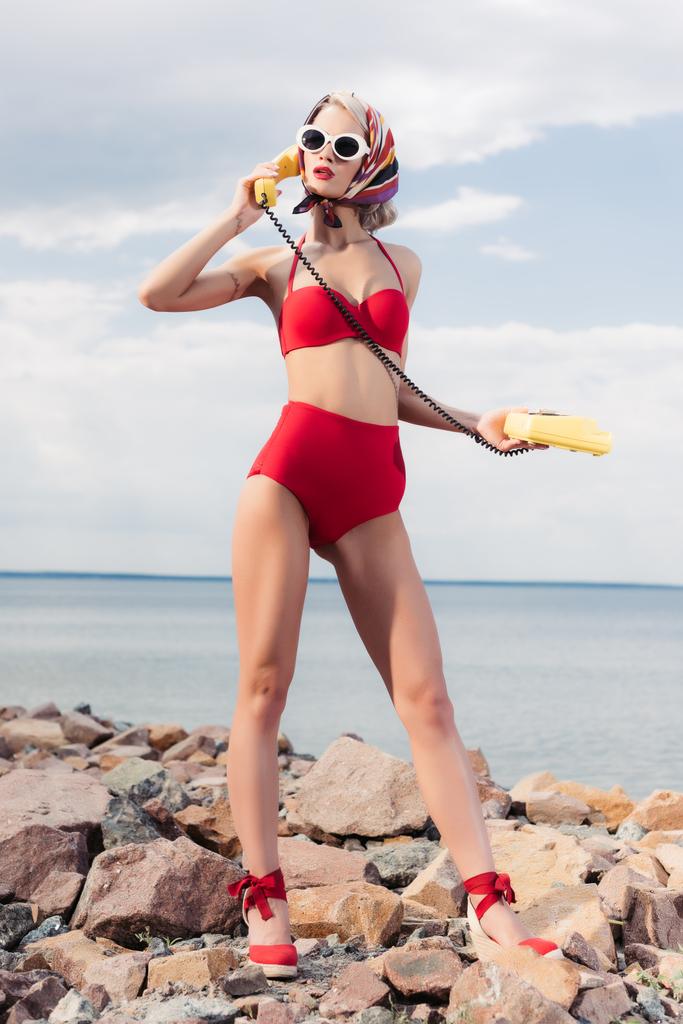 stylish woman in red bikini and silk scarf talking on yellow rotary phone and standing on shore - Photo, Image