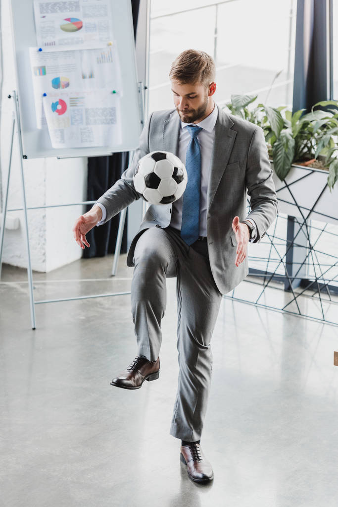 Ganzkörperansicht eines jungen Geschäftsmannes in formeller Kleidung, der im Büro mit Fußball spielt - Foto, Bild