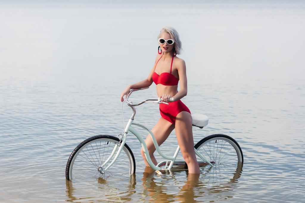 элегантная девушка в красном винтажном бикини позирует с велосипедом в воде
 - Фото, изображение
