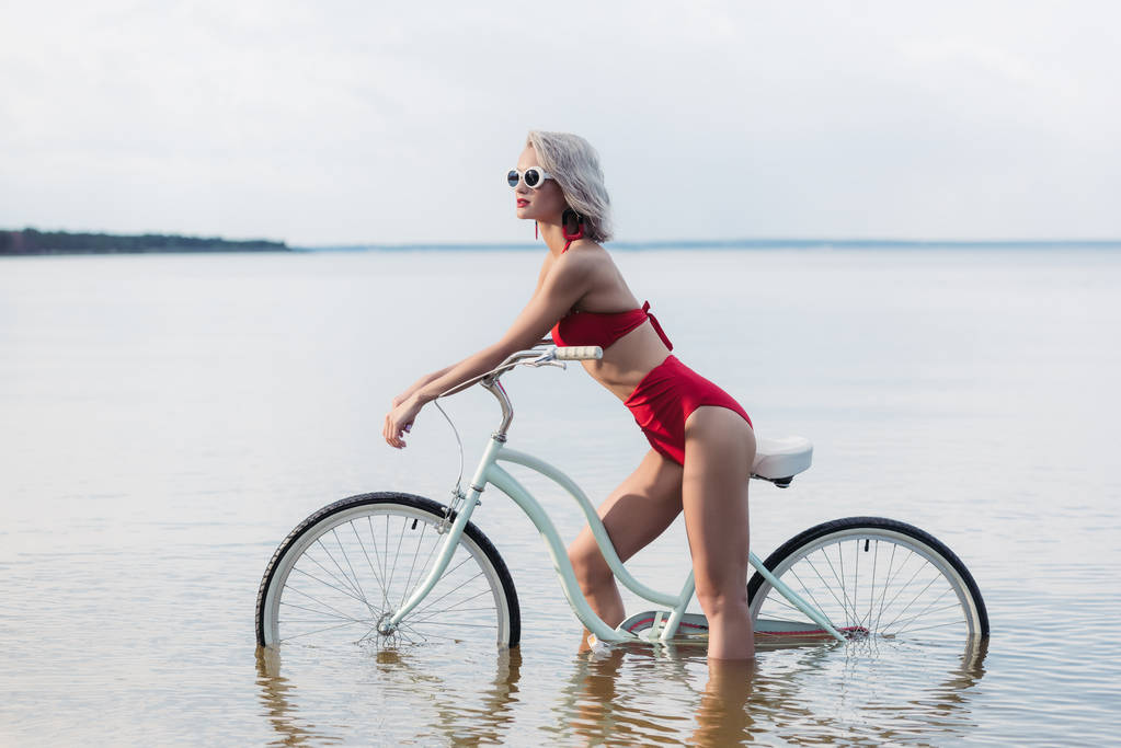 όμορφο κορίτσι της μόδας σε κόκκινο μπικίνι, κάθονται σε ποδήλατο στο νερό - Φωτογραφία, εικόνα