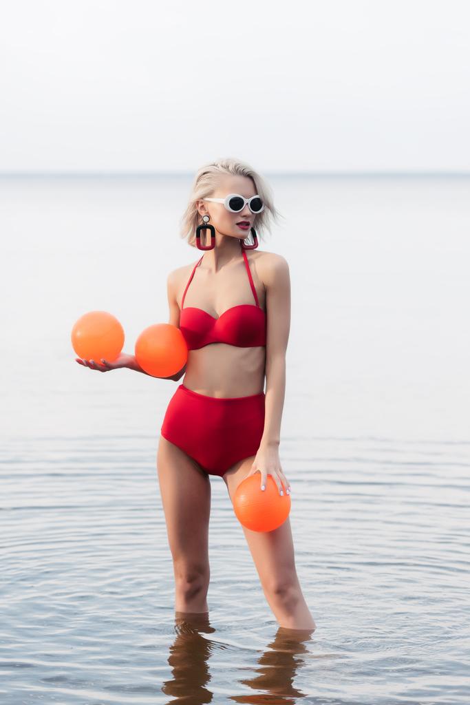 menina na moda em biquíni vermelho vintage e óculos de sol posando na água com bolas de laranja
 - Foto, Imagem
