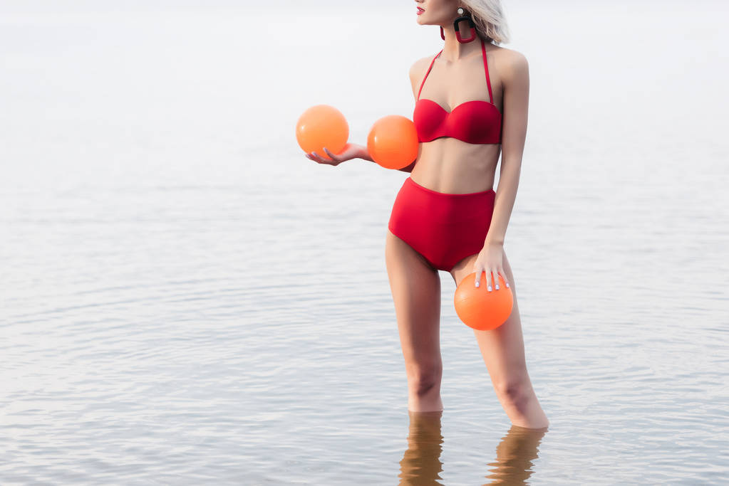 Ausgeschnittene Ansicht eines stilvollen Mädchens im roten Bikini, das im Wasser steht und orangefarbene Kugeln in der Hand hält - Foto, Bild