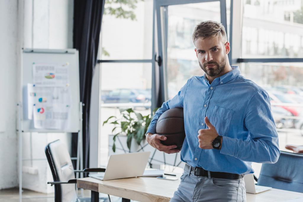 όμορφος νέος επιχειρηματίας που κρατά μπάλα μπάσκετ και δείχνει τον αντίχειρα στο γραφείο - Φωτογραφία, εικόνα