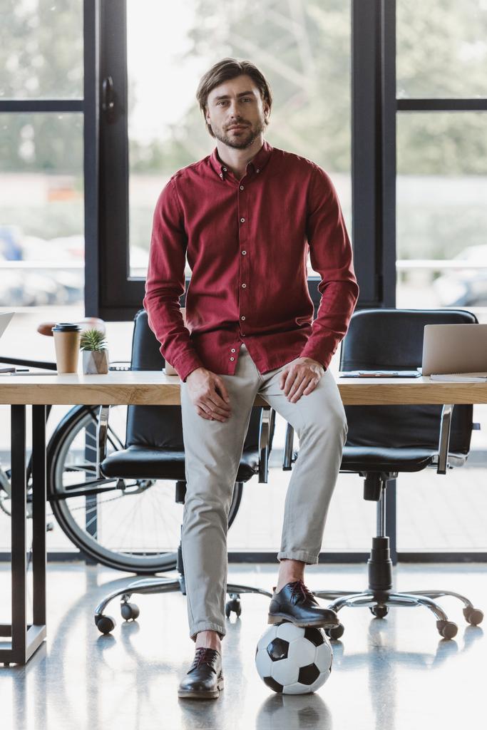 knappe jonge zakenman met voetbal zittend op tafel en kijken naar camera in kantoor - Foto, afbeelding