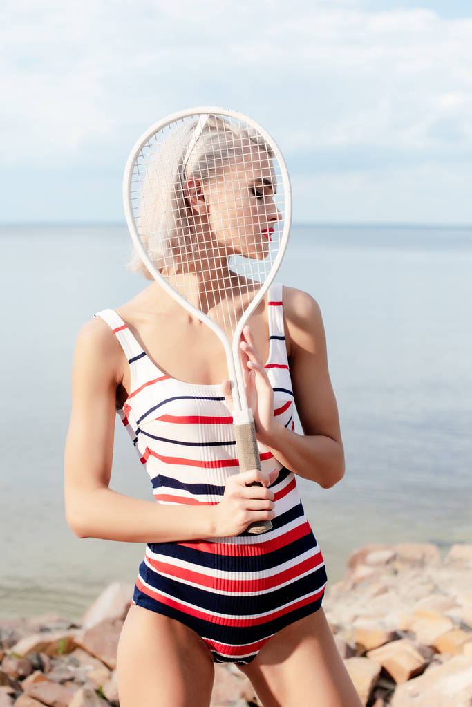 Blondynka atrakcyjna kobieta w kostium kąpielowy w paski pozowanie z tenisową na brzegu - Zdjęcie, obraz