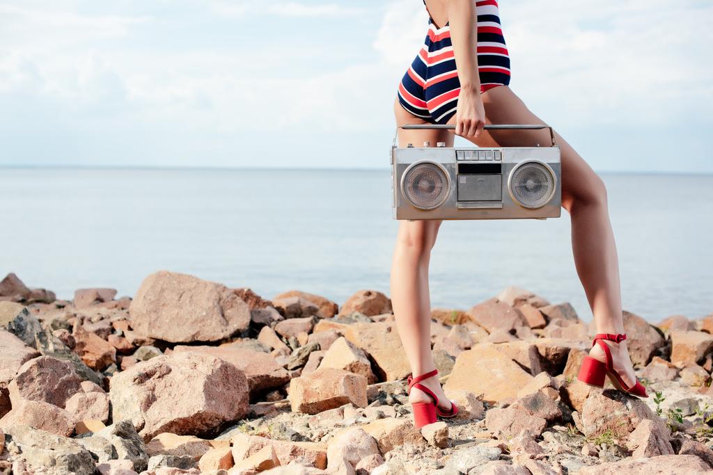cropped view of woman in swimsuit holding vintage boombox on rocks near the sea - Photo, Image