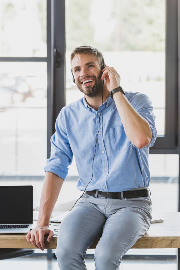 knappe lachende jonge call center-operator in hoofdtelefoon zittend op tafel in kantoor - Foto, afbeelding
