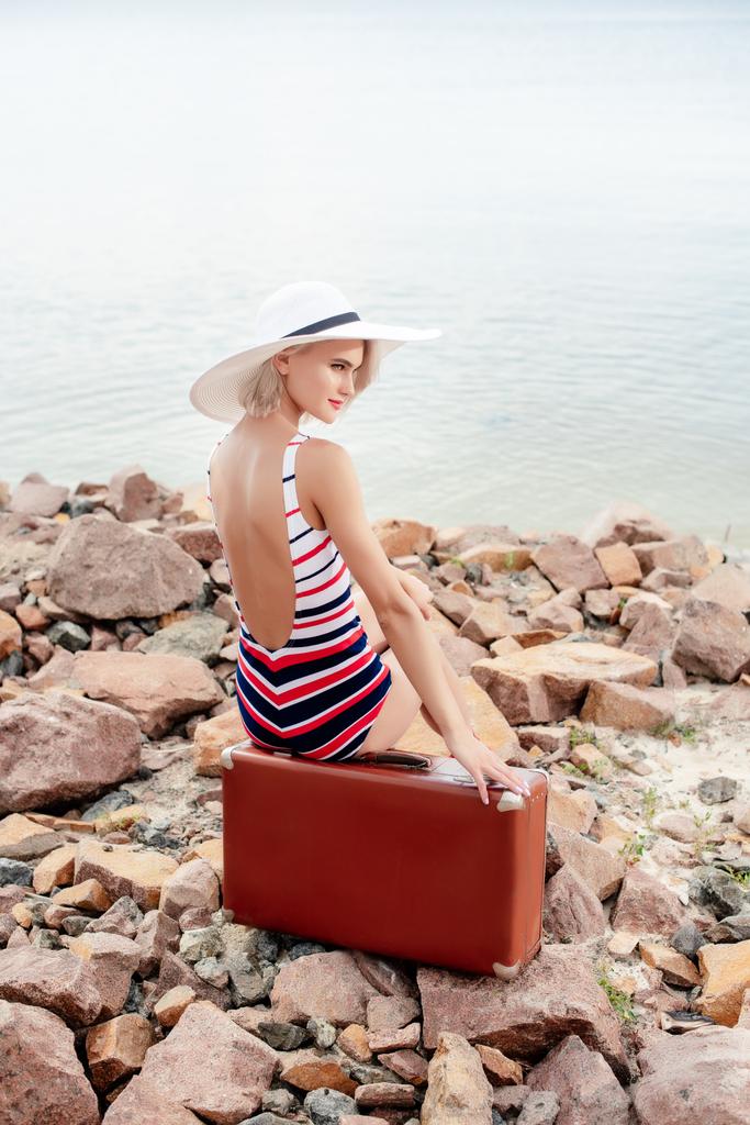 menina atraente em maiô vintage sentado no saco de viagem na praia rochosa
 - Foto, Imagem