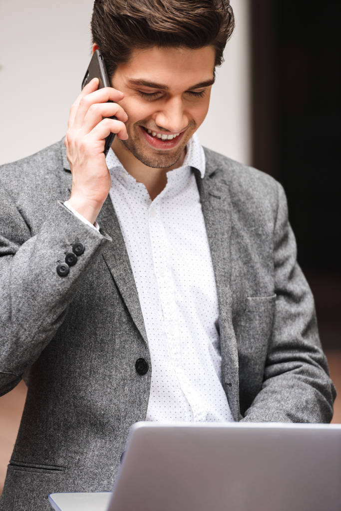 Vrolijke jonge zakenman gekleed in pak praten op mobiele telefoon terwijl u werkt aan laptop computer zitten buiten - Foto, afbeelding