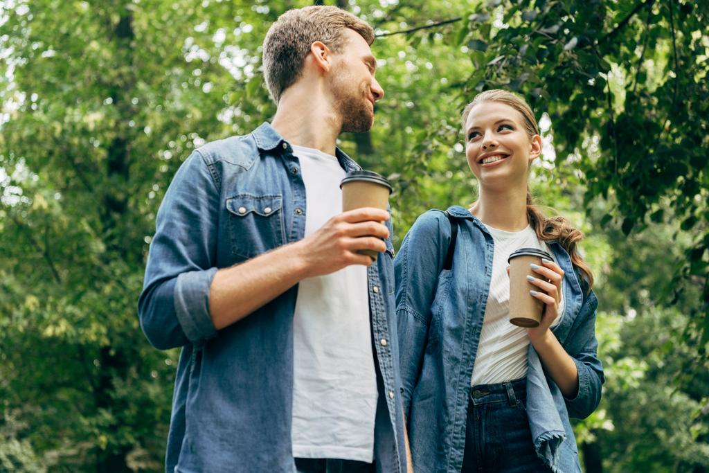 Blick von unten auf ein schönes junges Paar mit Pappbechern Kaffee, das gemeinsam im Park spaziert - Foto, Bild