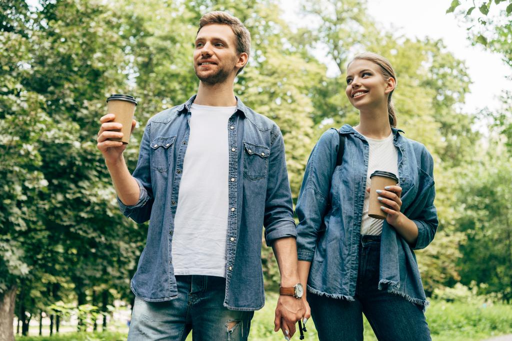 vista inferior de la feliz pareja joven con tazas de papel de café caminando por el parque juntos
 - Foto, Imagen
