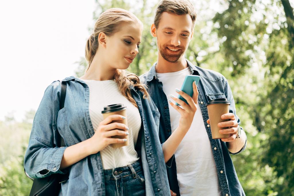 красива молода пара з паперовими чашками використовує смартфон разом у парку
 - Фото, зображення