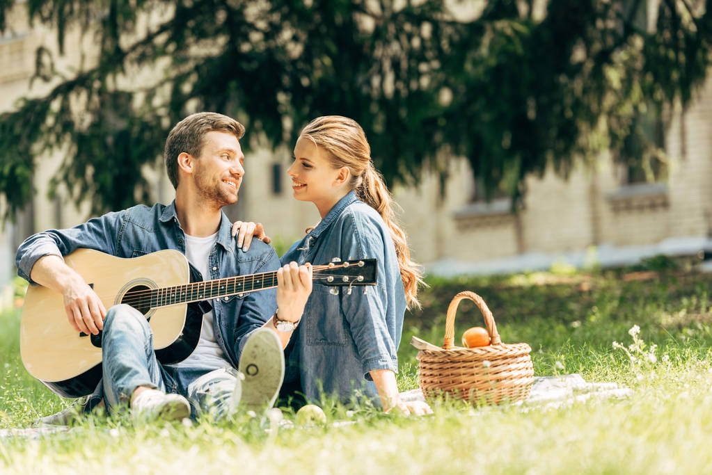 atractivo joven tocando la guitarra para su novia sonriente durante el picnic en el parque
 - Foto, Imagen