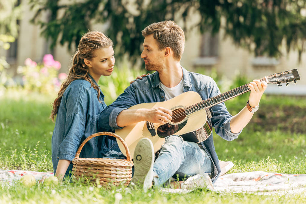 jovem bonito tocando guitarra para sua namorada sorridente durante piquenique no parque
 - Foto, Imagem