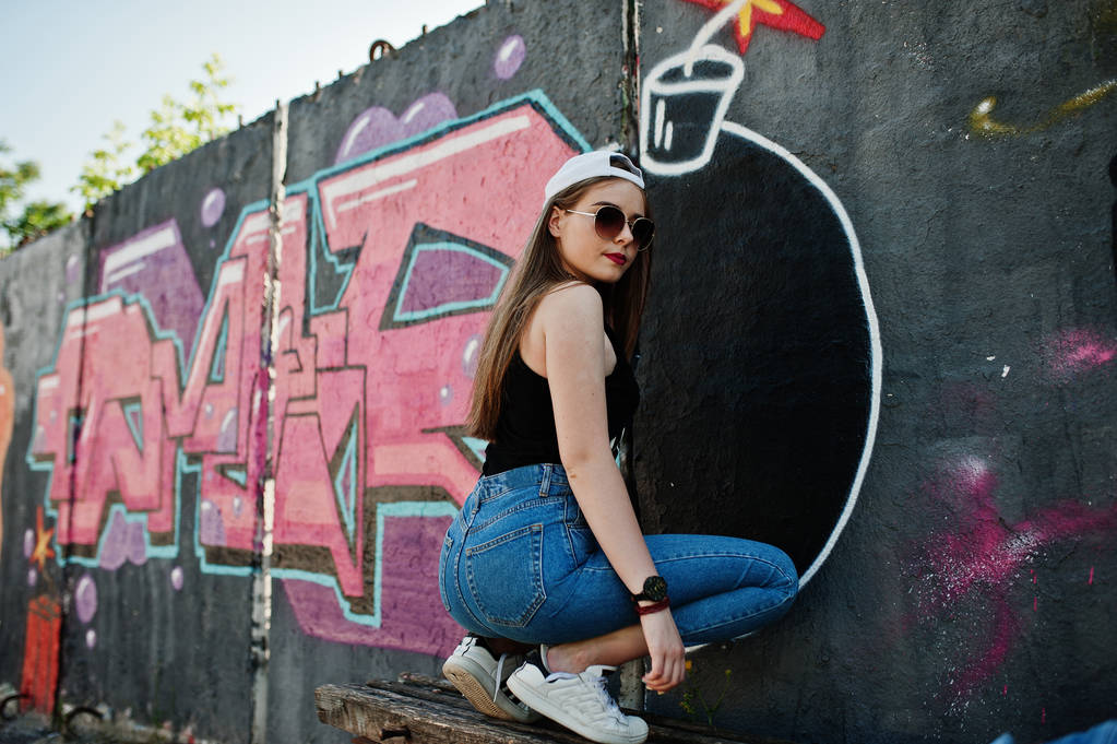 Indietro di natiche elegante ragazza hipster casual in berretto, occhiali da sole e jeans abbigliamento, ascoltando musica da cuffie del telefono cellulare contro grande parete graffiti con grande tnt bomba
. - Foto, immagini