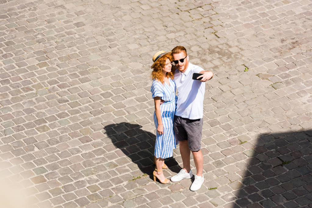 hoge hoekmening van roodharige man in zonnebril selfie met vriendin op smartphone bij stad straat - Foto, afbeelding