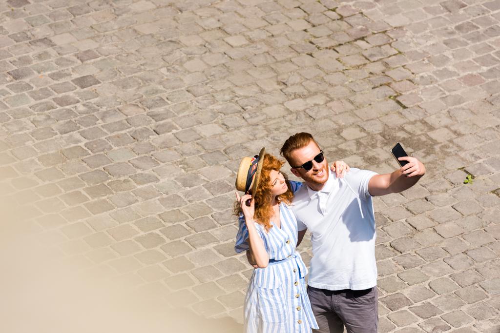 high angle view of smiling redhead man in sunglasses taking selfie with girlfriend on smartphone at city street - Photo, Image