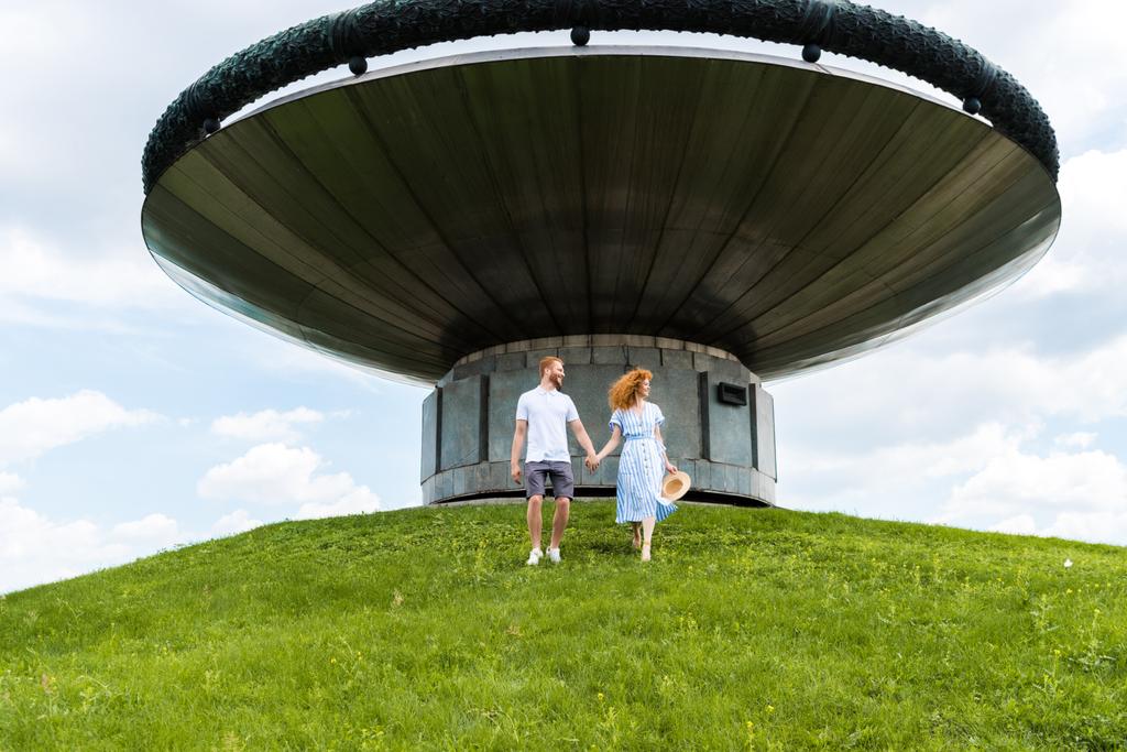 pelirroja pareja cogida de la mano y caminando en la colina cubierta de hierba en frente de un edificio moderno
  - Foto, Imagen