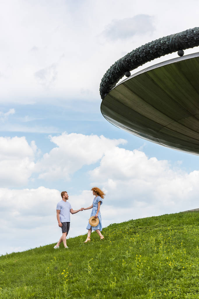 vista distante della coppia rossa che si tiene per mano sulla collina erbosa vicino all'edificio contro il cielo nuvoloso blu
 - Foto, immagini