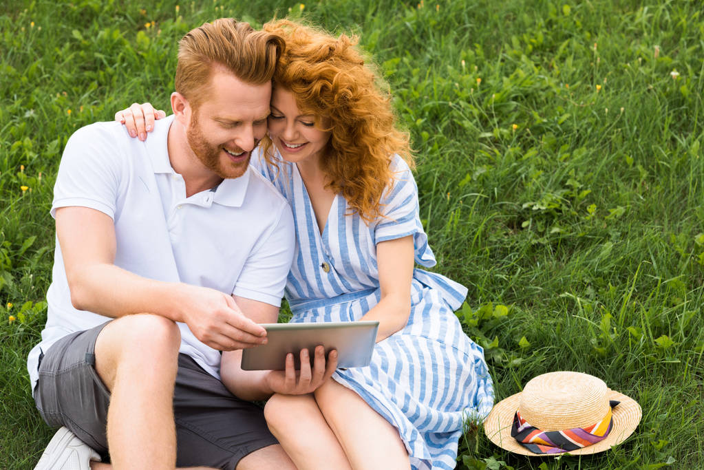 улыбающаяся рыжая пара с цифровой таблеткой на травянистом лугу
 - Фото, изображение