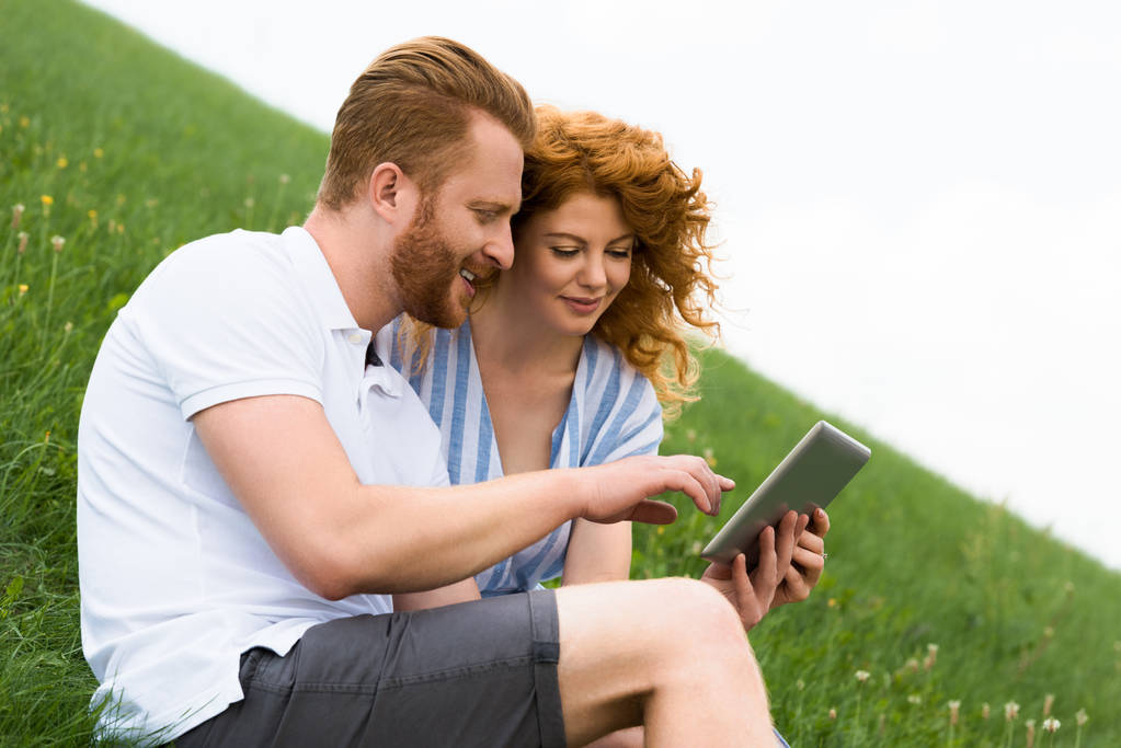 Zijaanzicht van het glimlachen roodharige man met digitale tablet terwijl zijn vriendin zit in de buurt van op de met gras begroeide heuvel  - Foto, afbeelding