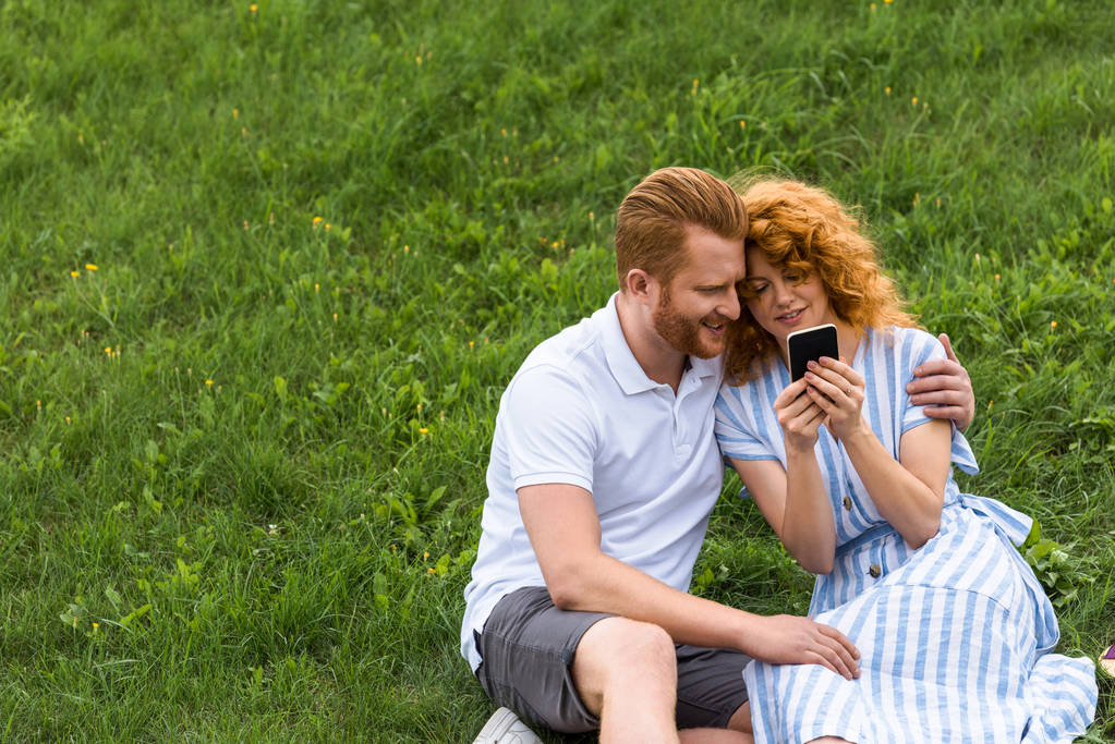 guapo pelirrojo hombre abrazando novia mientras ella mostrando él smartphone en verde prado
  - Foto, Imagen