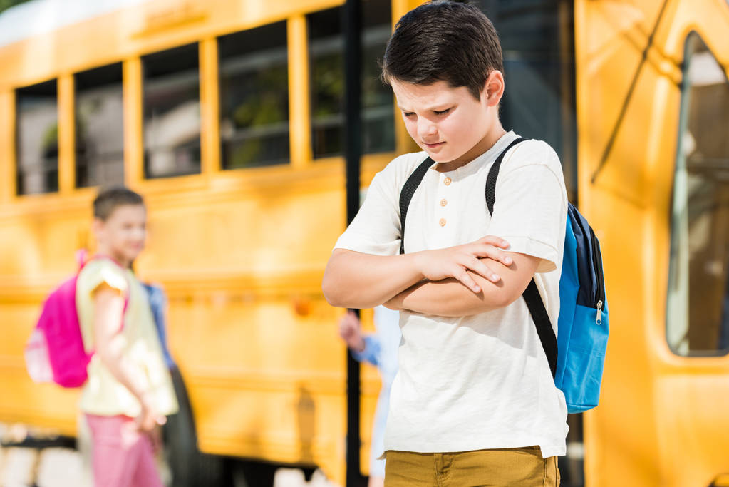πιεσμένη μικρή μαθητής με σταυρωμένα χέρια που στέκεται μπροστά από το σχολικό λεωφορείο - Φωτογραφία, εικόνα