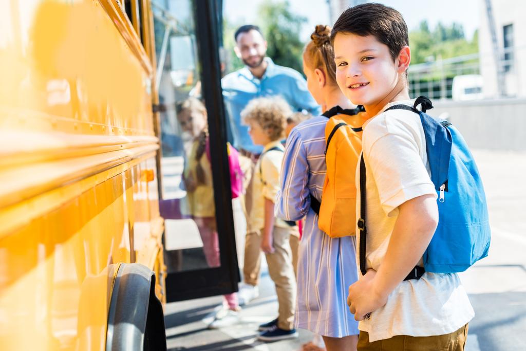 little schoolboy entering school bus with classmates while teacher standing near door - Photo, Image