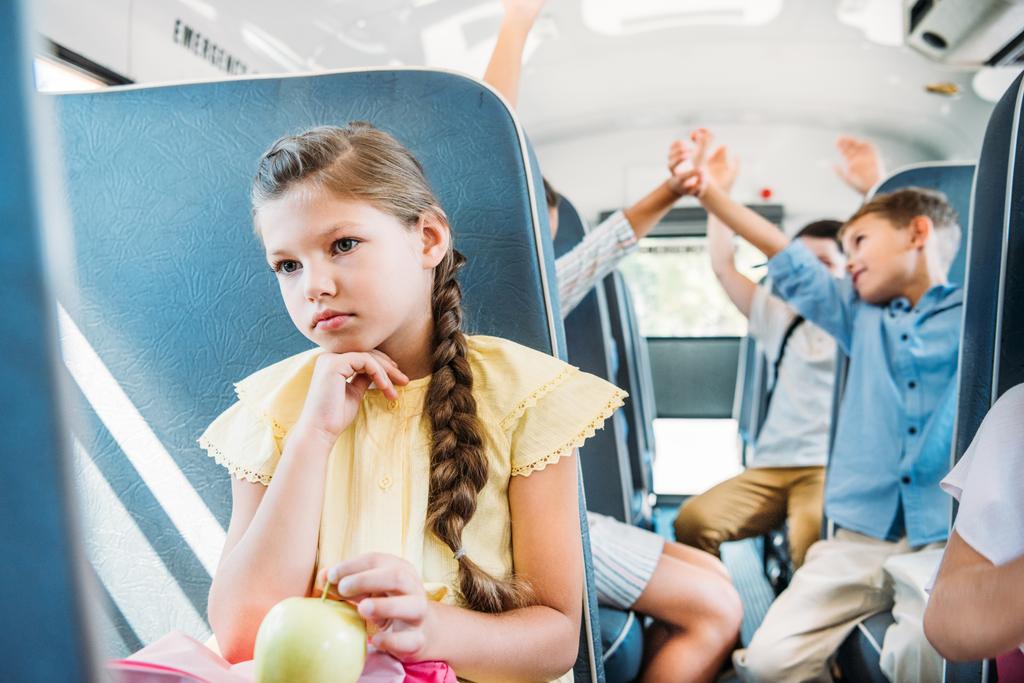 triest weinig schoolmeisje rijden op schoolbus terwijl haar klasgenoten hoog geven vijf vaag op de achtergrond - Foto, afbeelding