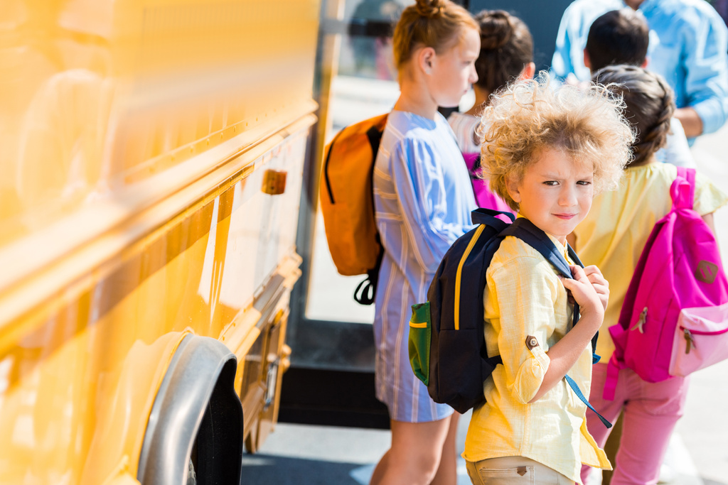 adorável colegial encaracolado com seus colegas de classe de pé perto do ônibus escolar
 - Foto, Imagem