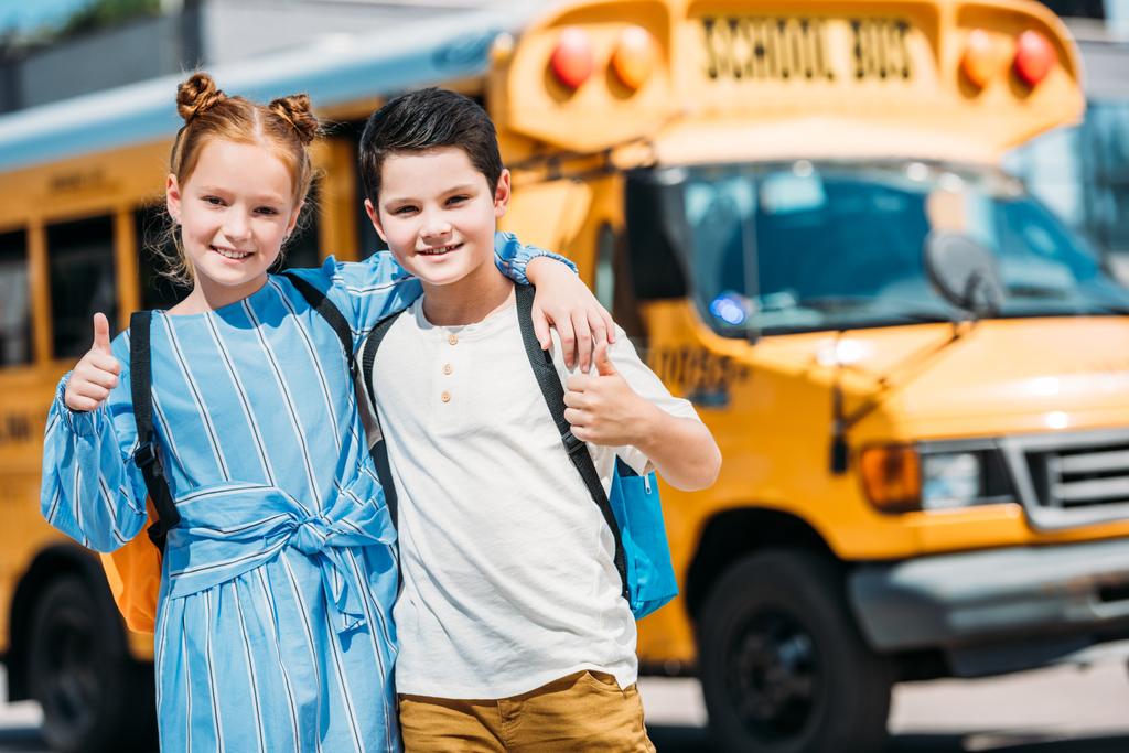счастливые маленькие ученые, показывающие большие пальцы и смотрящие в камеру перед школьным автобусом
 - Фото, изображение