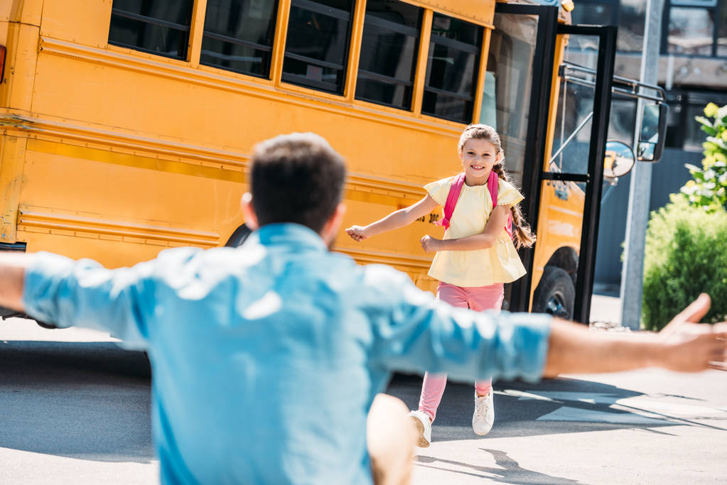 Отец с распростертыми объятиями ждет дочь, пока она бежит из школьного автобуса
 - Фото, изображение