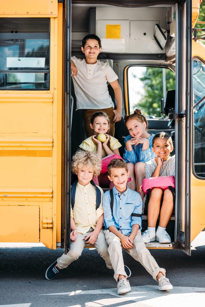 group of cute schoolchildren sitting on stairs of school bus and looking at camera - Photo, Image