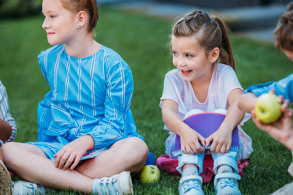 adorable little schoolgirls sitting on grass and laughing together - Photo, Image