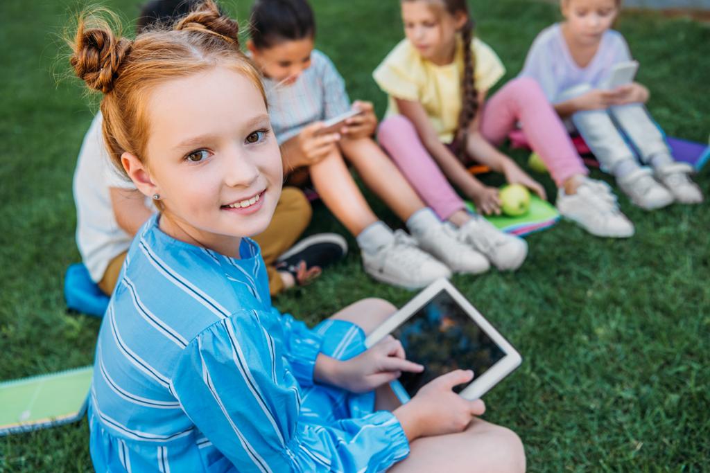 sourire adorable écolière en utilisant la tablette tout en étant assis sur l'herbe avec des camarades de classe
 - Photo, image