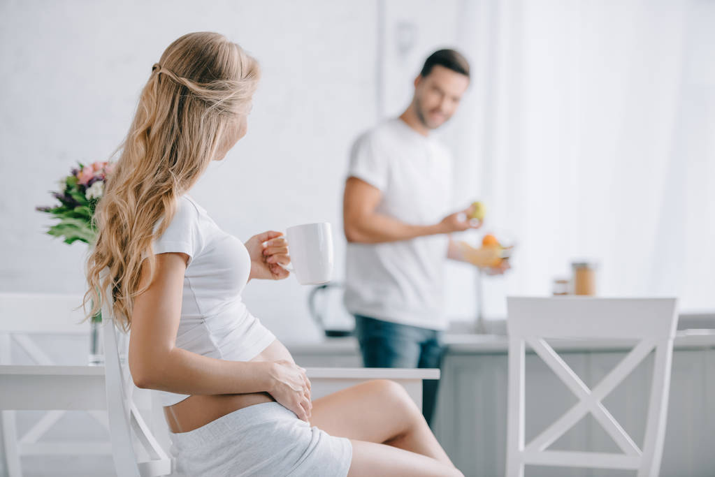 enfoque selectivo de la mujer embarazada con taza de té sentado en la silla y el marido de pie en el mostrador en la cocina
 - Foto, imagen