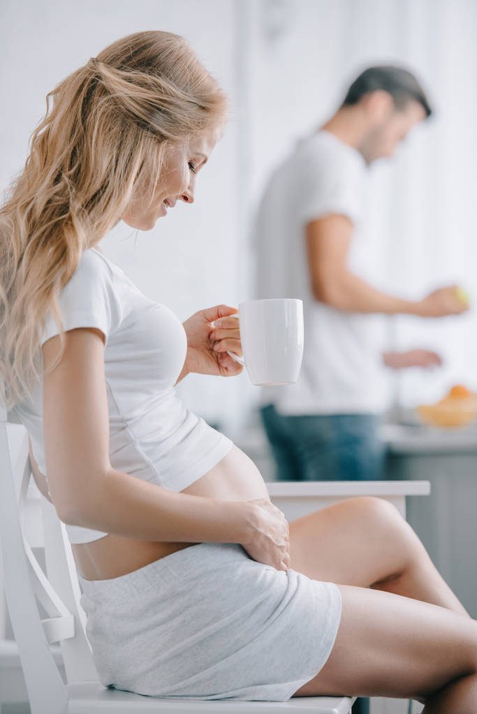 επιλεκτική εστίαση της εγκύου με φλυτζάνι του τσαγιού που κάθεται σε καρέκλα και σύζυγος στέκεται στο πάγκο στην κουζίνα - Φωτογραφία, εικόνα