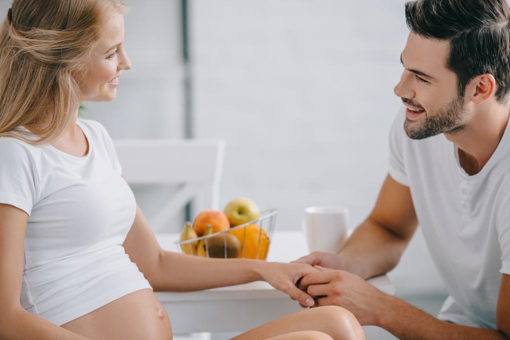 sivunäkymä hymyilevä raskaana oleva nainen ja aviomies kädestä pöydässä kotona
 - Valokuva, kuva