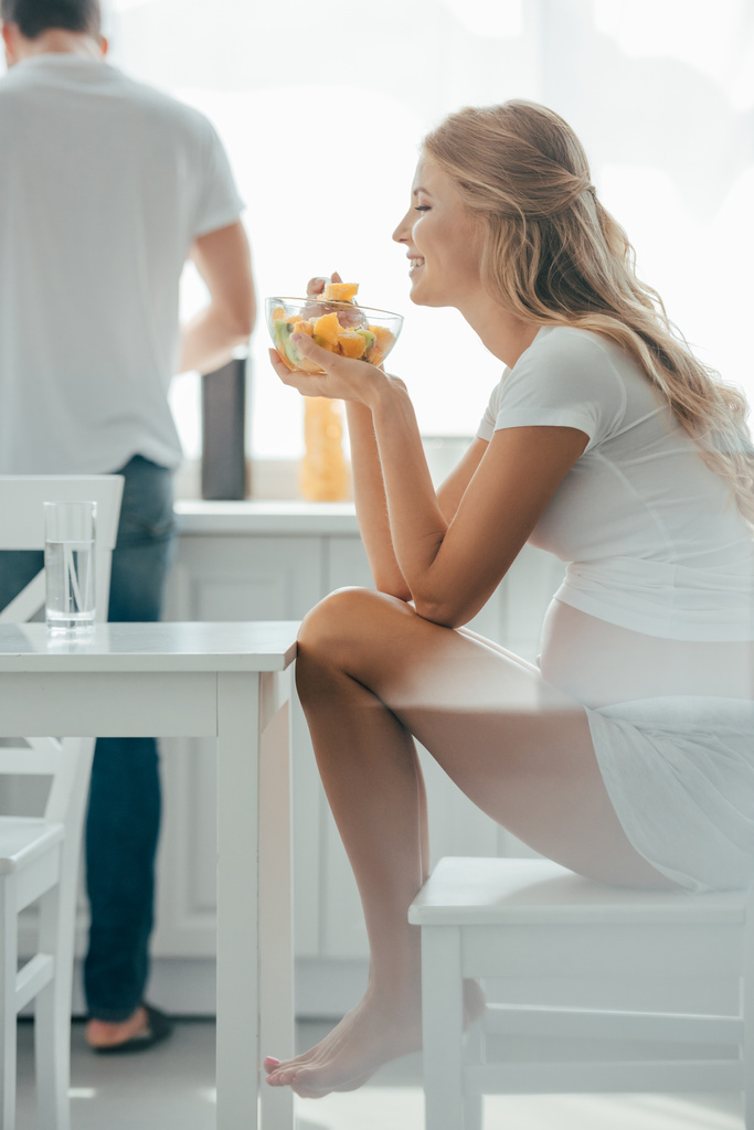 selective focus of smiling pregnant woman eating fruits salad while husband standing at counter in kitchen - Photo, Image