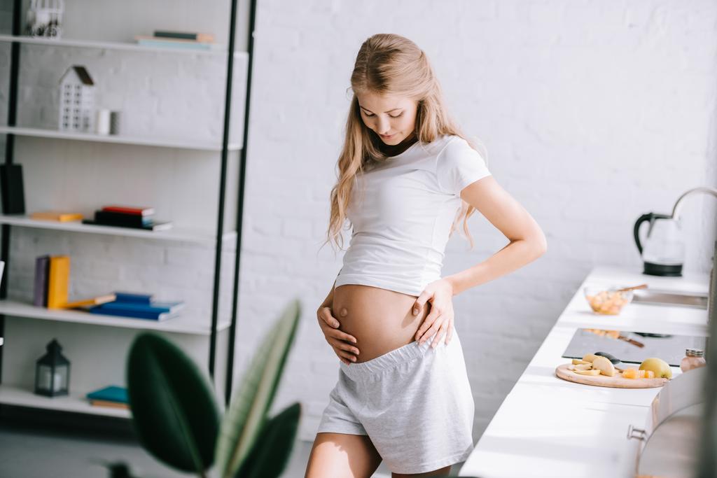 красива вагітна жінка з руками в животі, стоячи вдома
 - Фото, зображення
