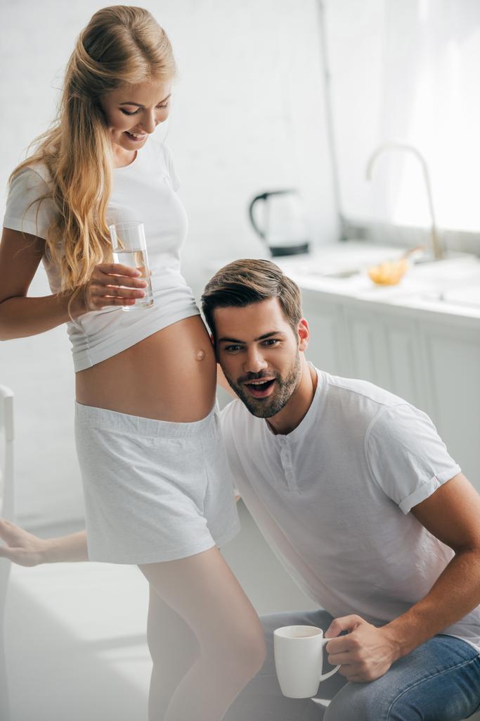 muž s šálkem kávy poslechu břicho těhotné manželky v kuchyni doma - Fotografie, Obrázek