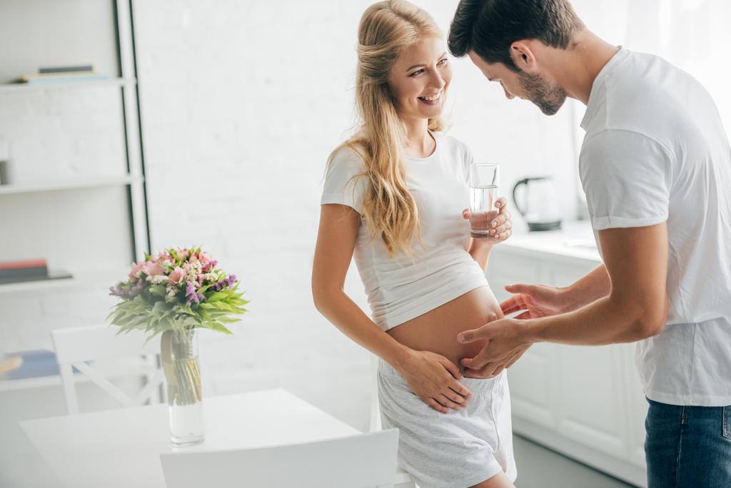husband touching belly of happy pregnant wife with glass of water at home - Photo, Image