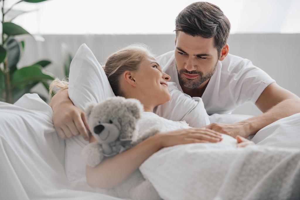 беременная женщина в белой ночнушке с плюшевым мишкой на диване и мужем рядом дома
 - Фото, изображение