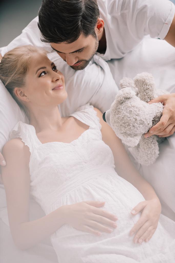 oyuncak ayı ve evde kanepe üzerinde beyaz Geceliği gülümseyen hamile karısı olan adam portresi - Fotoğraf, Görsel