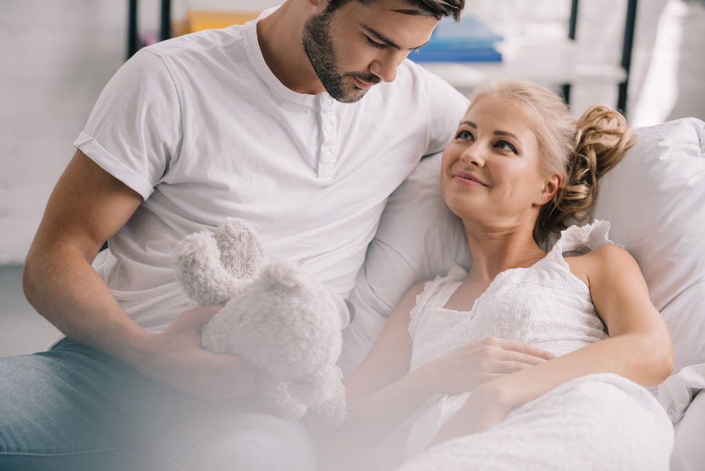 portrait of man with teddy bear sitting near pregnant wife in white nightie on sofa at home - Photo, Image