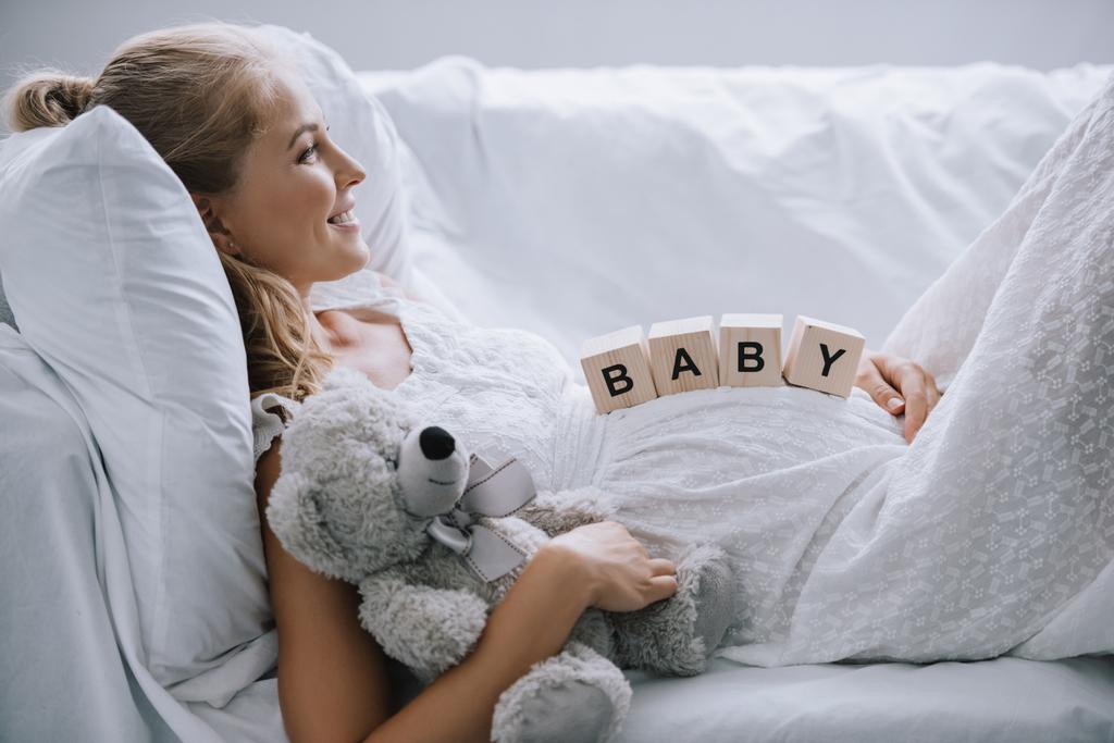 Seitenansicht einer lächelnden Schwangeren im weißen Nachthemd mit Teddybär und Holzblöcken mit Babyaufdruck auf dem Sofa - Foto, Bild