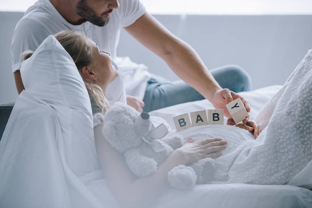 partial view of man putting wooden blocks with baby lettering on belly of pregnant wife with teddy bear at home - Photo, Image