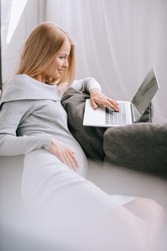 вид сбоку беременной женщины, использующей ноутбук на диване дома
 - Фото, изображение