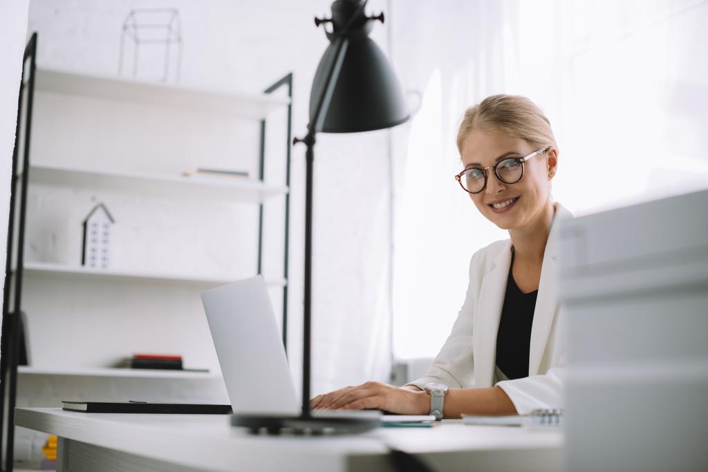 портрет улыбающейся деловой женщины в очках, смотрящей в камеру на рабочем месте с ноутбуком в офисе
 - Фото, изображение