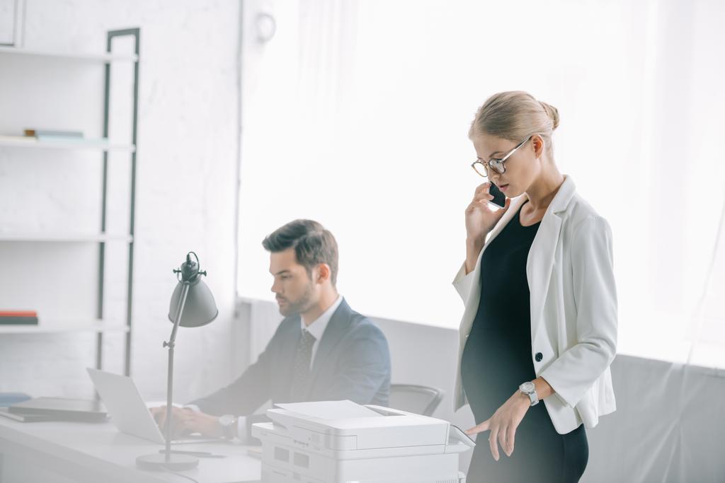 Schwangere Geschäftsfrau spricht auf Smartphone, während Kollegin am Laptop am Arbeitsplatz im Büro arbeitet - Foto, Bild